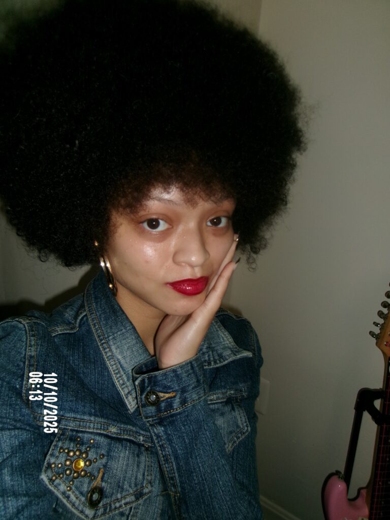 A selfie of a girl with an afro smiling while her hand is on her chin in a cute way.