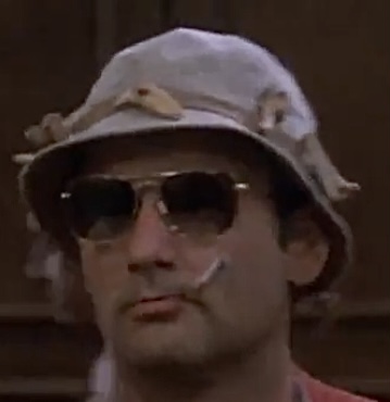 A disheveled Bill Murray wearing a boonie hat and dark shades, looking rather glum with a bullfrog like frown as long cigarette juts out of his mouth