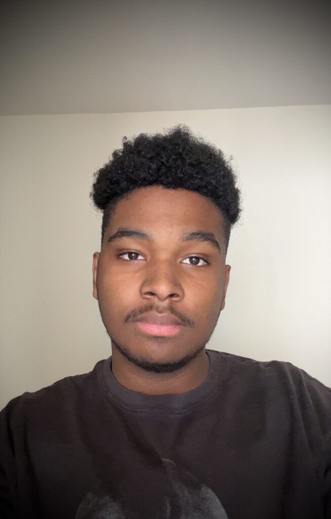 A person facing the camera indoors against a plain light-colored wall. They have a neutral expression on thier face, wearing a dark T-shirt